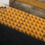 Modern bir bina girişinde, görme engelli bireyin beyaz bastonuyla dokunduğu, paslanmaz çelikten yapılmış Trimetal hissedilebilir takip ve uyarı yüzeyi.